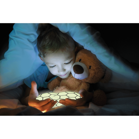 Ein Kind sitzt im Bett, hält ein leuchtendes Objekt in den Händen und umarmt einen Teddybären, während es unter der Decke ist.