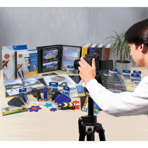 Eine Person fotografiert eine Vielzahl von Büro- und Bastelmaterialien, darunter Ordner, Kleber und bunte Formen, auf einem Tisch.