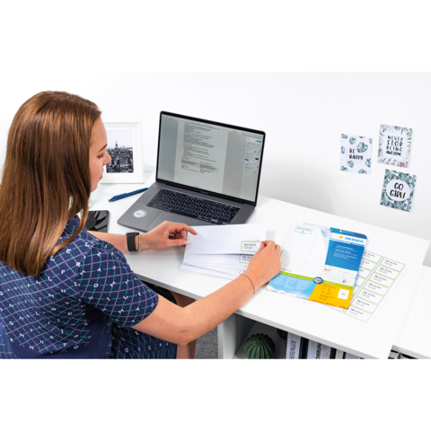 Eine Person sitzt an einem Schreibtisch, arbeitet am Laptop und sortiert Dokumente und Briefe. Auf dem Tisch liegen verschiedene Unterlagen.